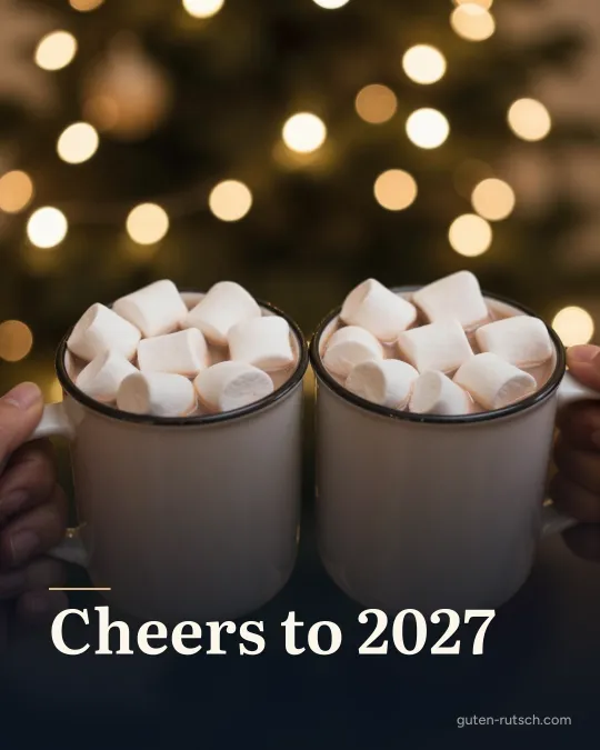 Zwei Tassen Kakao mit Marshmallows vor einer Lichterkette, darauf der Gruß „Cheers to 2027“